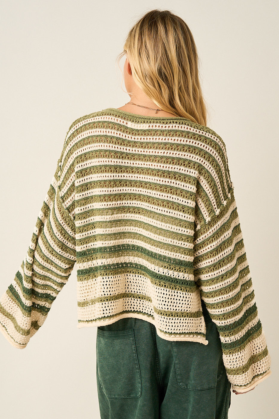 Back of women wearing a striped crochet boxy sweater in dark sage  and green pants.