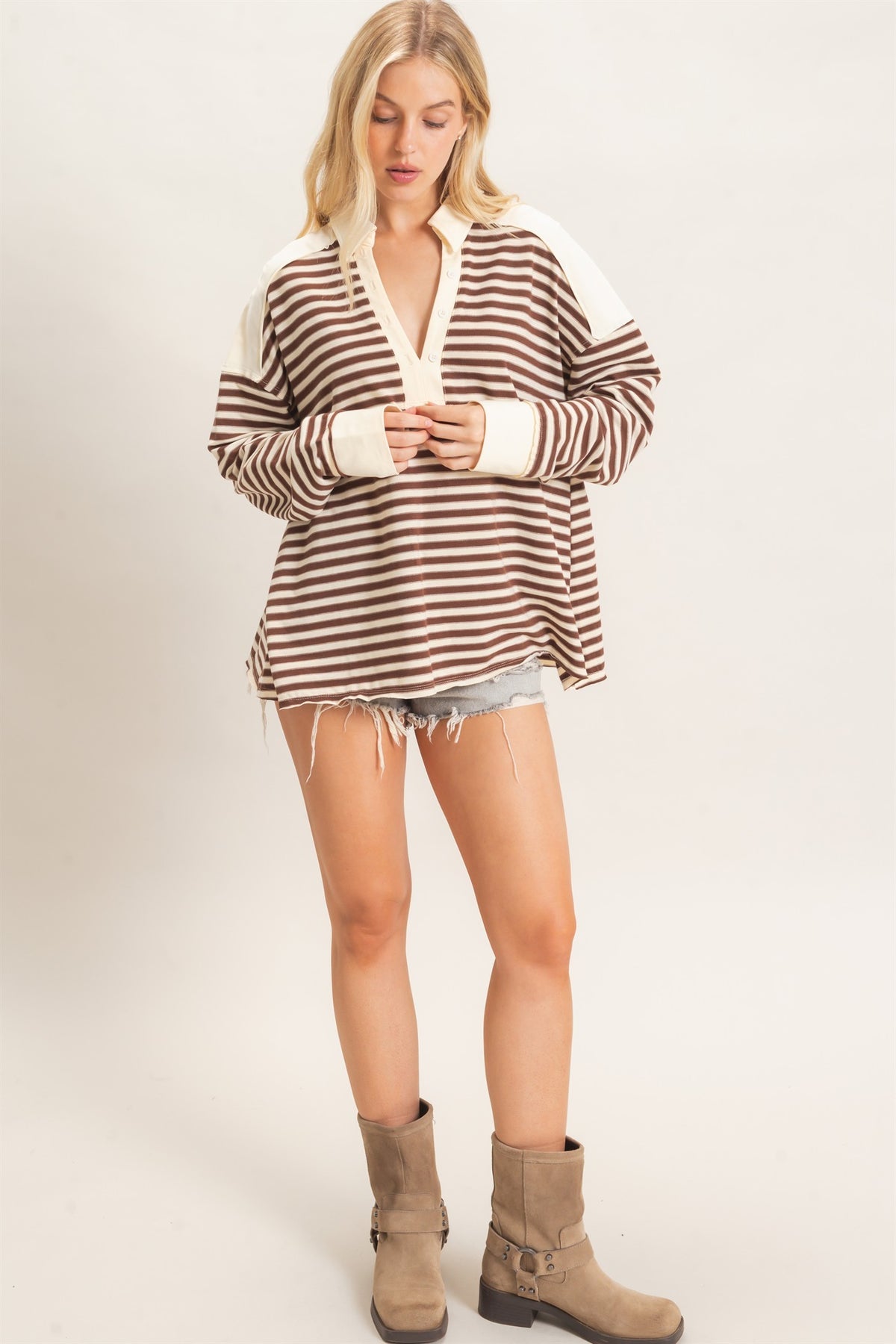A woman modeling a brown and cream striped long-sleeve henley sweatshirt with a V-neck and frayed hem, paired with denim shorts and taupe ankle boots, against a plain light background.