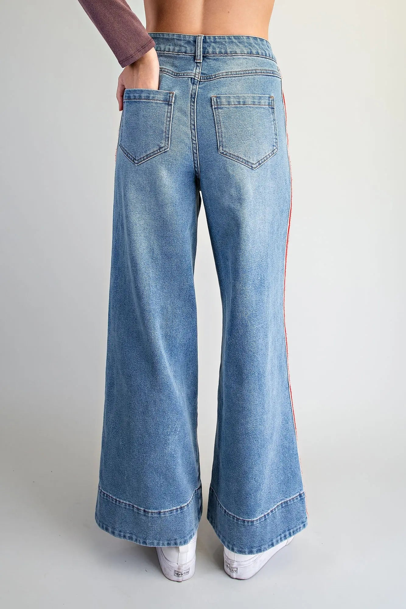 A person wearing light blue wide-leg denim jeans with a red and white vertical stripe down the side, paired with white sneakers, shown against a plain light gray studio background.