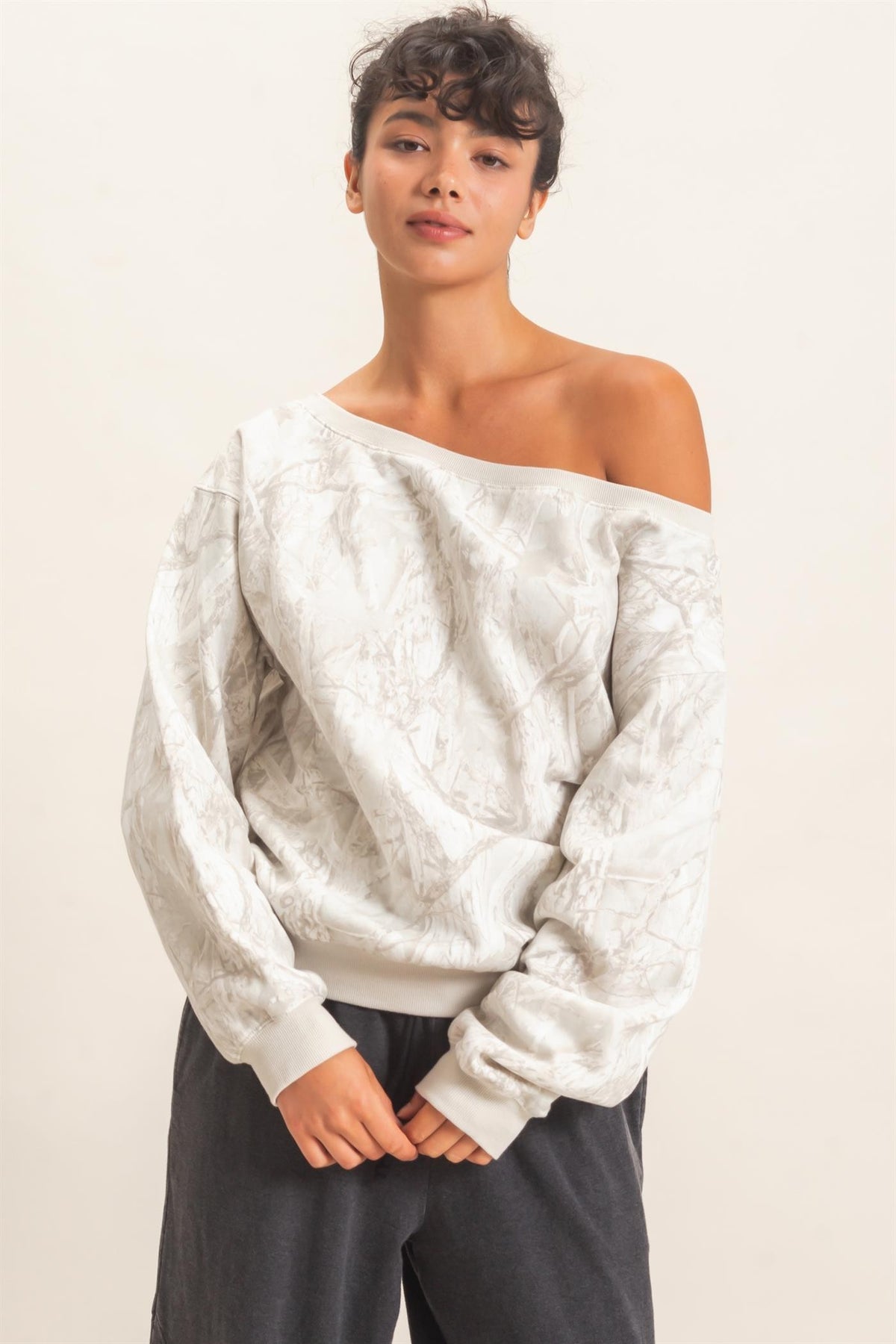 A woman modeling a cream-colored off-the-shoulder knit sweater with a subtle tonal tree camo pattern, paired with dark gray pants, against a plain off-white background.