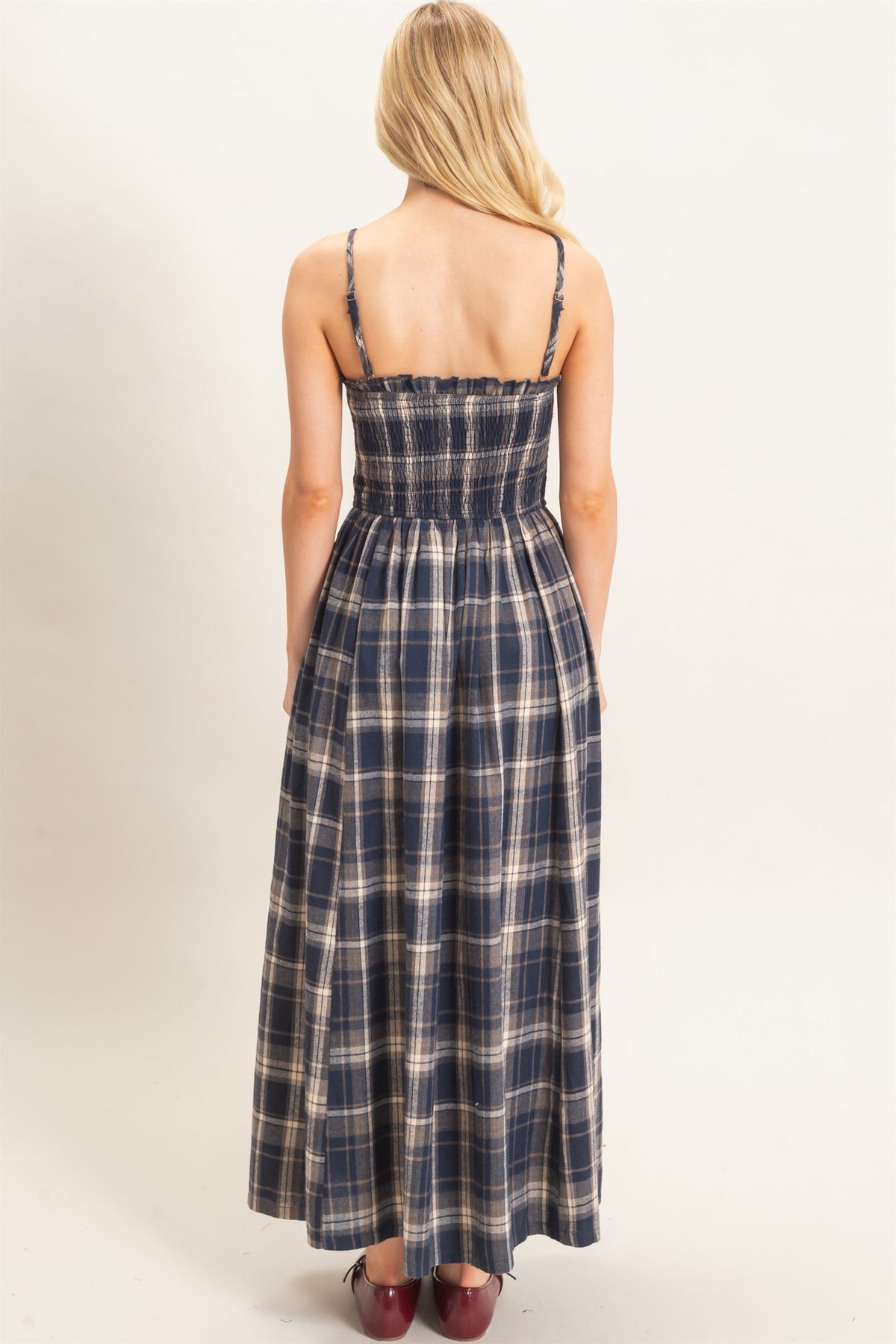 A woman viewed from the back wearing a sleeveless plaid maxi dress with thin straps and a smocked bodice, paired with dark red shoes, against a plain light background.