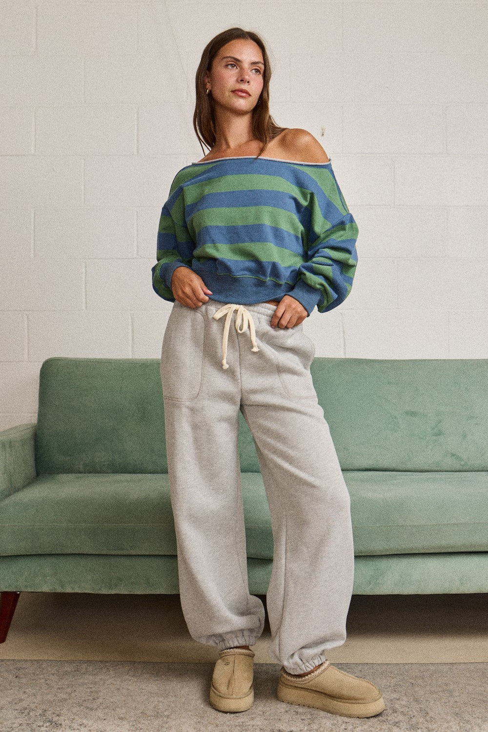 A woman stands in front of a green couch wearing a blue and green striped off-the-shoulder sweater, light gray drawstring sweatpants, and tan slip-on shoes.