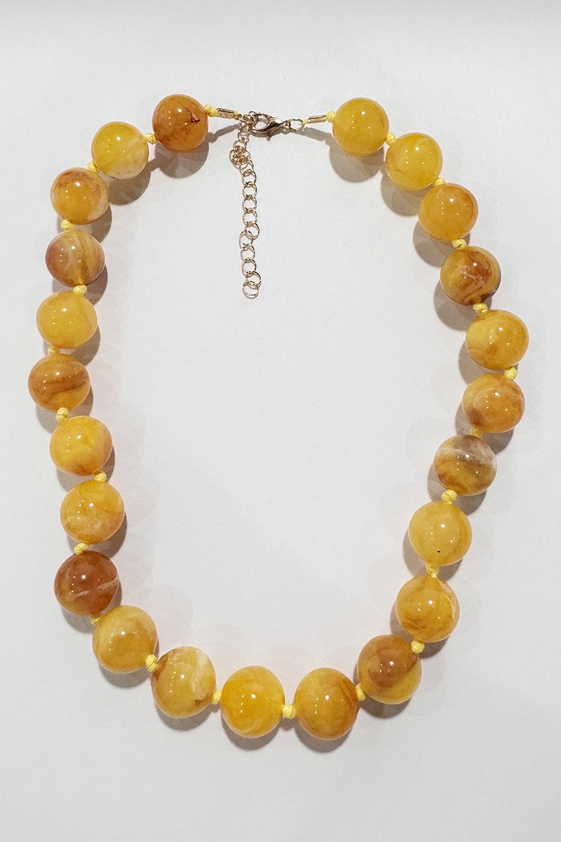 A beaded necklace made of round, polished yellow-orange stone beads with natural marbling and some translucent qualities, connected by a gold-tone chain extender and clasp, displayed on a light off-white background.