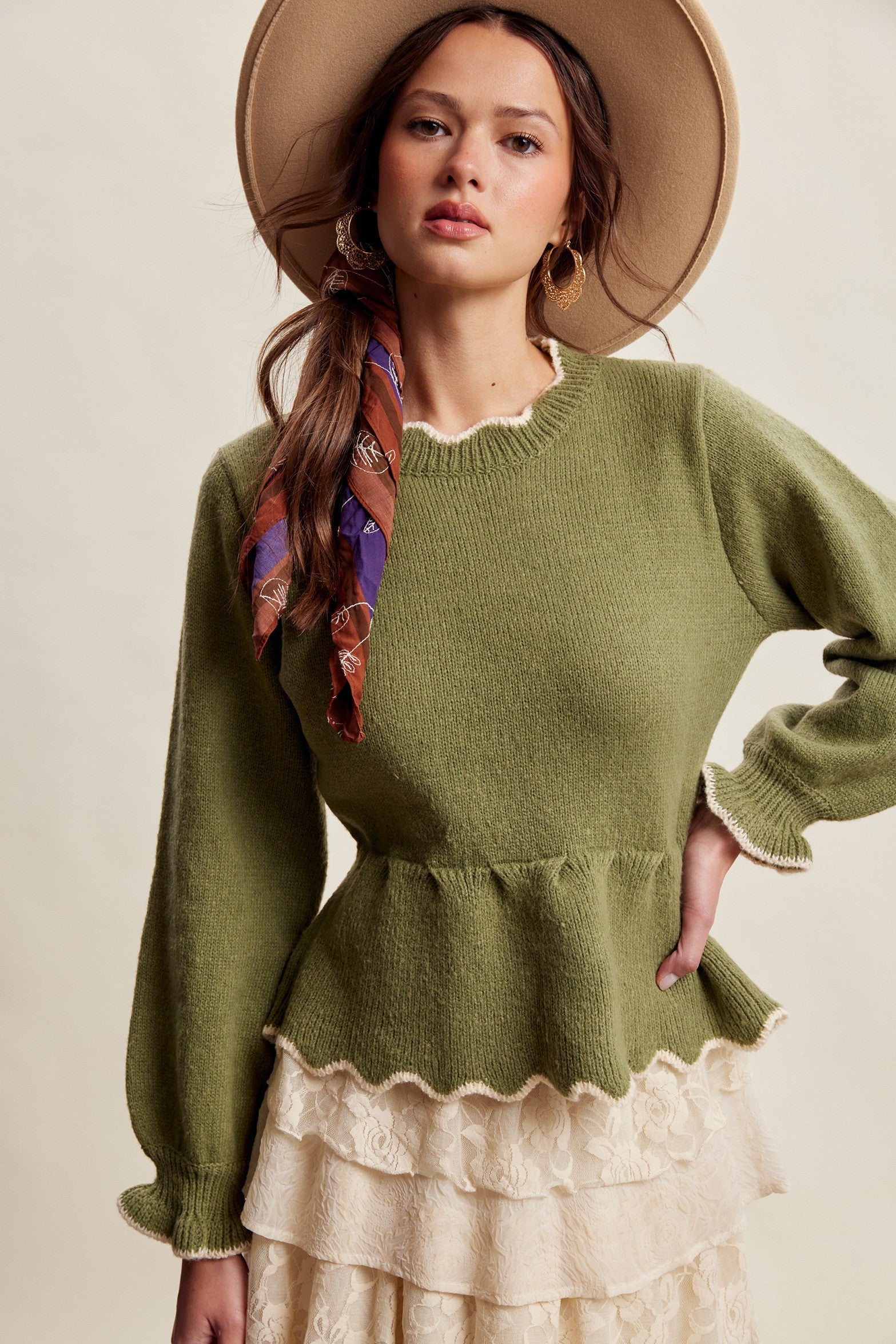A woman wearing an olive green knit sweater with a ruffled peplum hem, layered over a cream lace tiered skirt, and wearing a wide-brimmed tan hat with a patterned scarf.