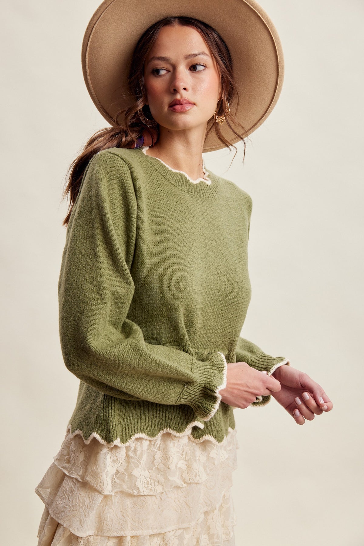 A woman wearing a olive green knit sweater with a scalloped neckline and cuffs, layered over a cream ruffled skirt and wearing a wide-brimmed tan hat, against a plain beige background.