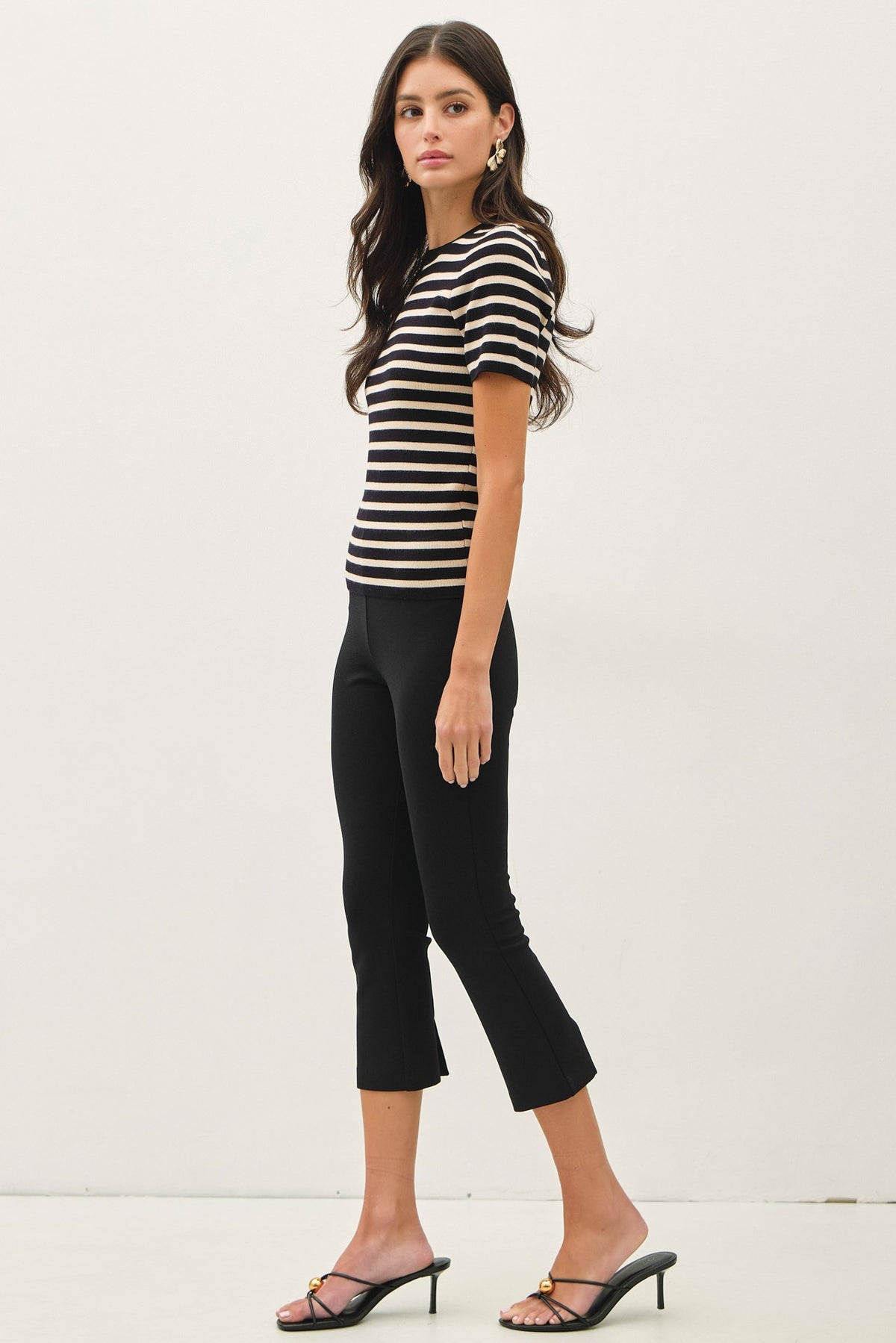 A woman modeling a short-sleeve black and white horizontally striped knit top paired with black cropped pants and black heeled slide sandals, photographed against a plain off-white studio background.