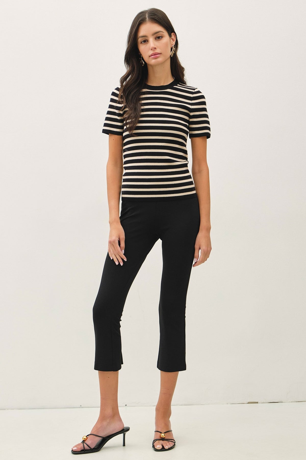 A woman modeling a short-sleeve black and beige striped t-shirt paired with black cropped pants and black strappy heels, standing against a plain white studio background.