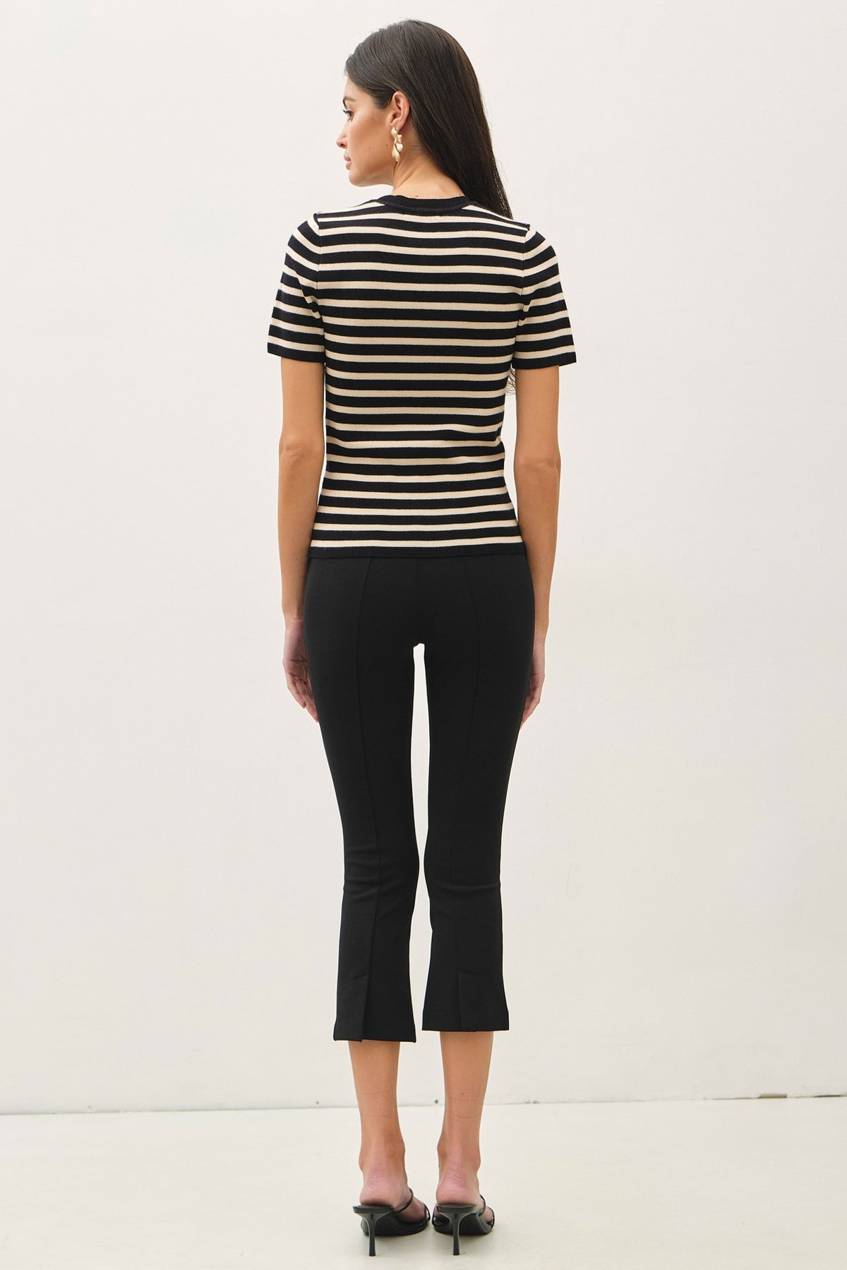 A woman shown from the back wearing a short-sleeve black and beige striped top with black cropped pants and black heels, standing against a plain white studio background.