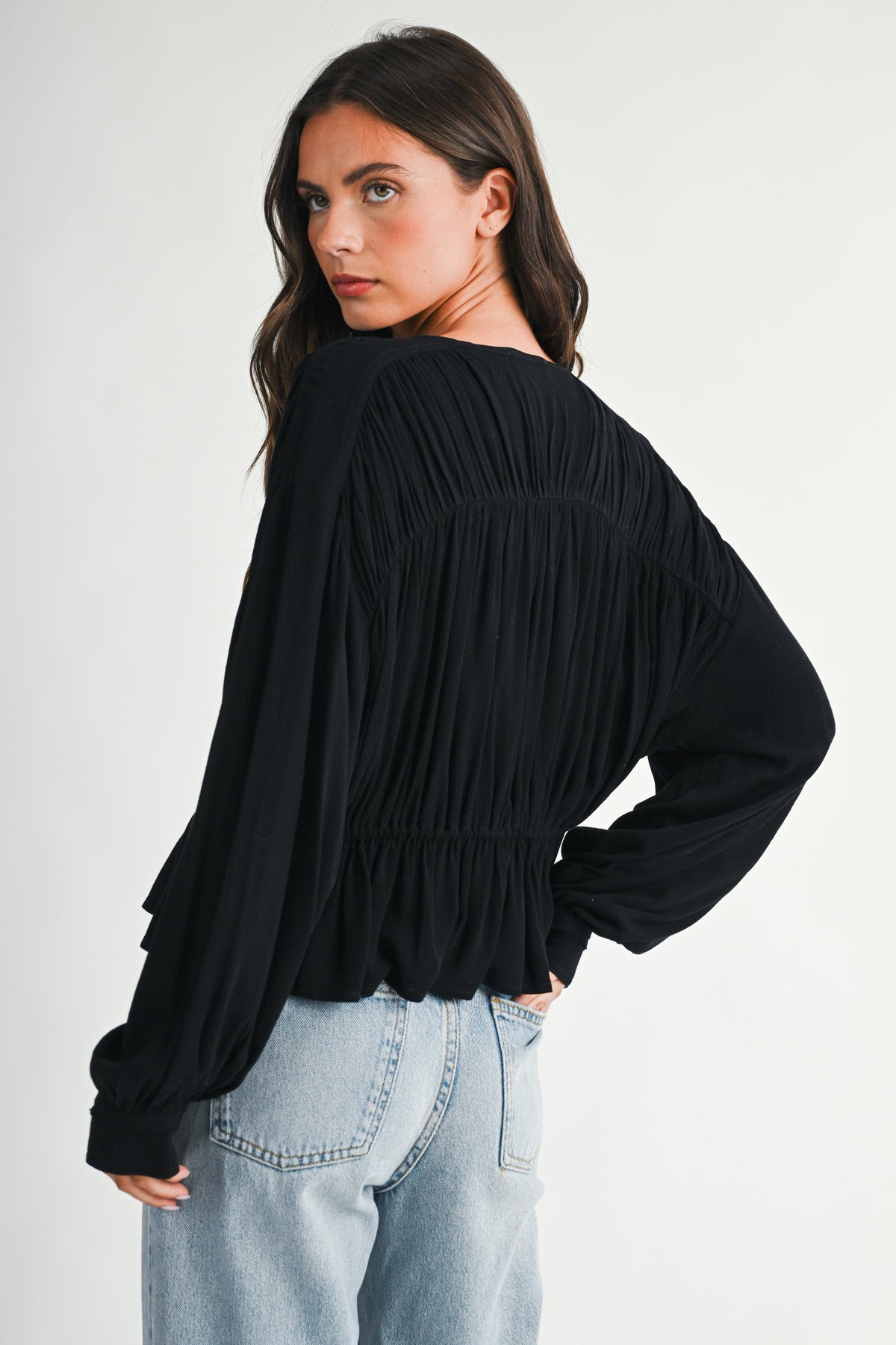 Dark haired women looking over her shoulder with textured peplum blouse in back and a pair of blue denim jeans on.