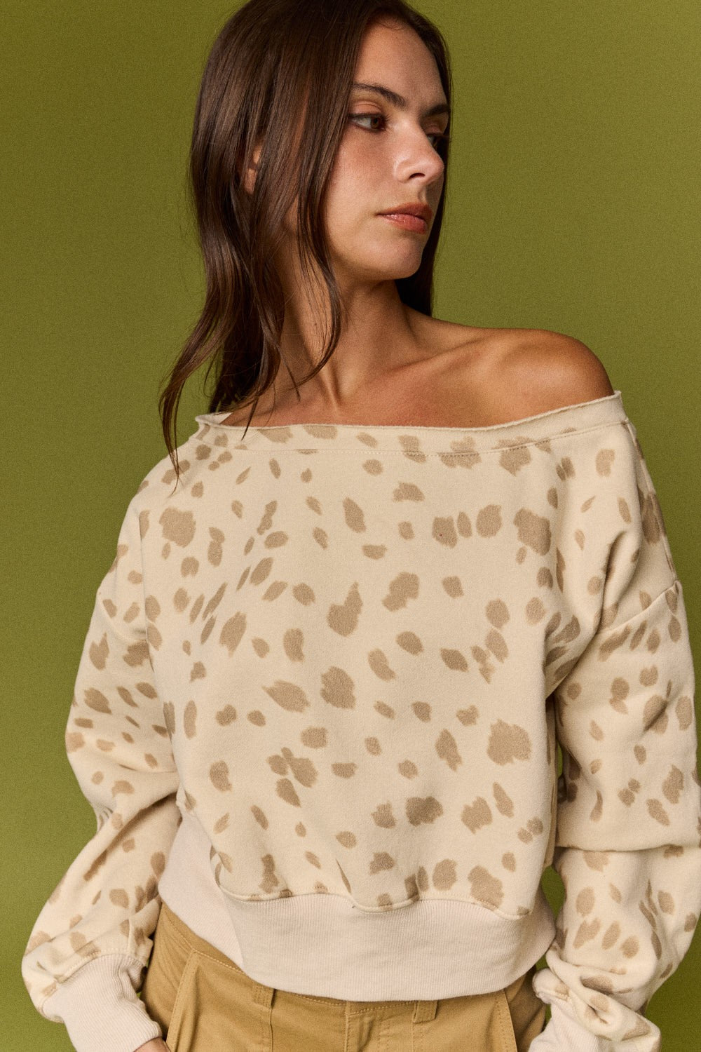 A woman wearing an off-the-shoulder beige and tan leopard-print sweatshirt, paired with khaki pants, set against a solid olive green background.