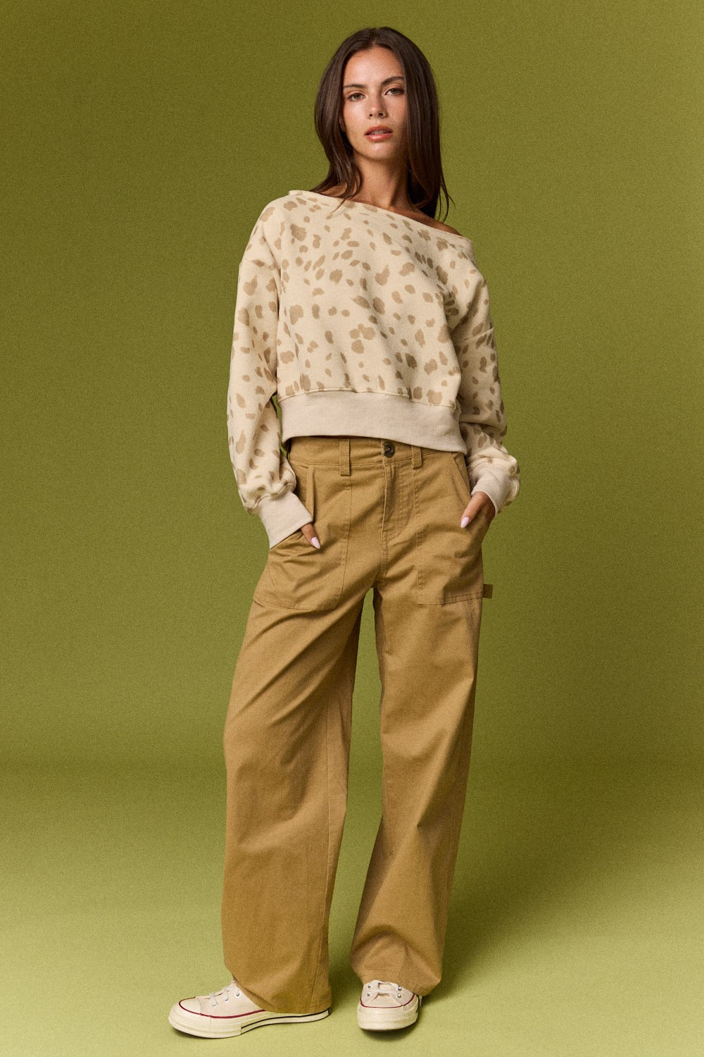 A model stands against a solid olive green studio backdrop wearing a beige and brown leopard-print off-the-shoulder sweater, wide-leg tan pants, and white sneakers.