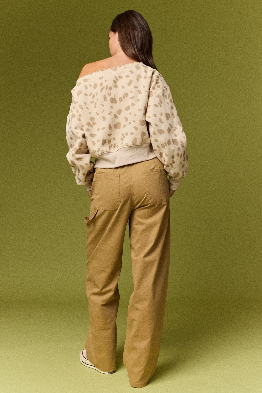 A model is shown from behind wearing a beige and brown animal print off-the-shoulder long-sleeve crop top and tan wide-leg cargo pants, set against a solid olive green studio background.