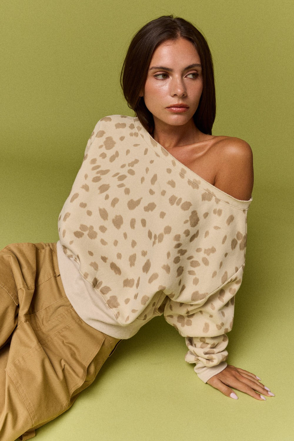 A woman wearing a beige and brown leopard-print off-the-shoulder long-sleeve top and tan pants, posing against a solid olive green background.