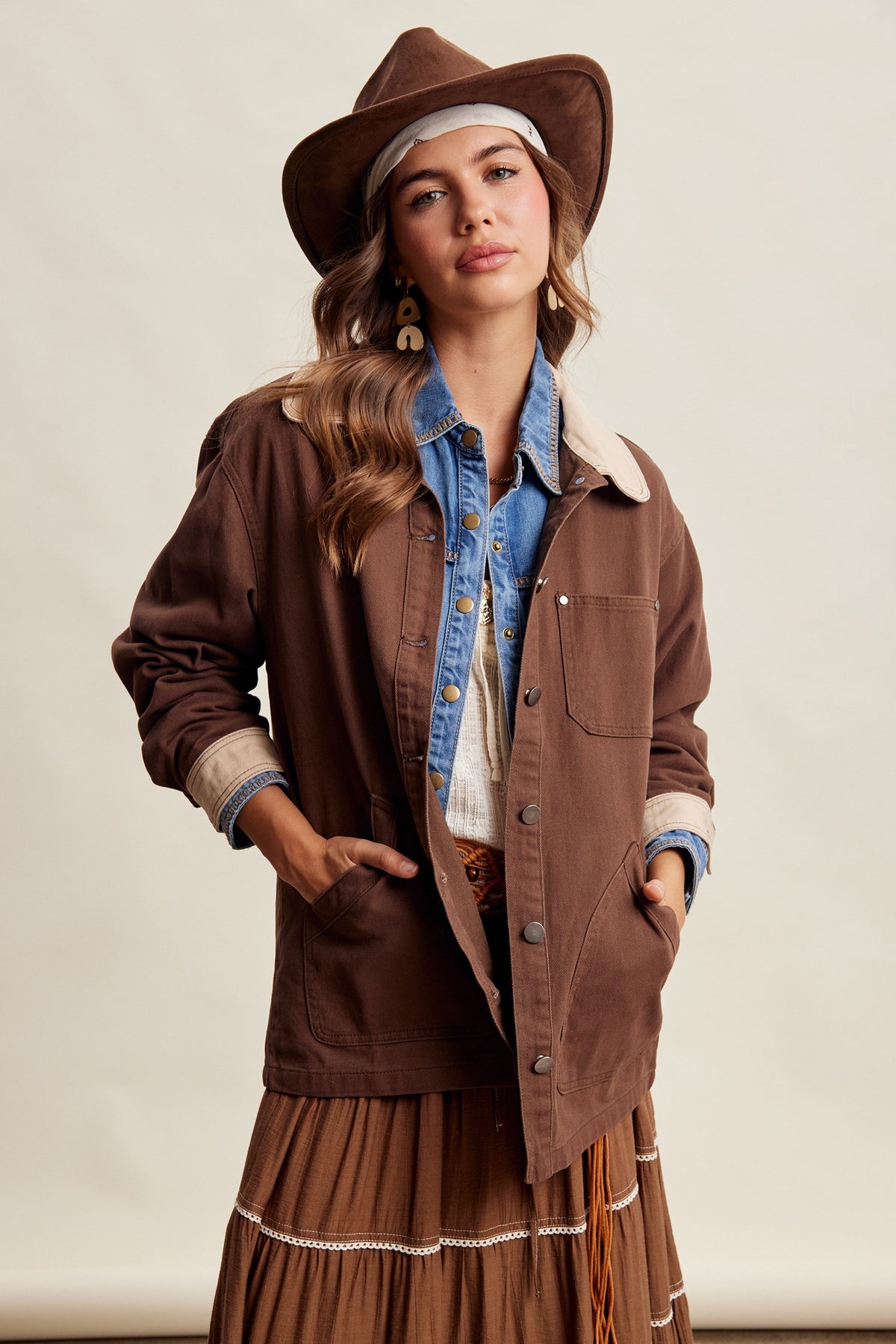 A woman modeling a brown espresso colored western-style utility jacket with a light brown shearling collar, worn over a blue denim shirt and a brown fringe skirt, complete with a matching brown cowboy hat.