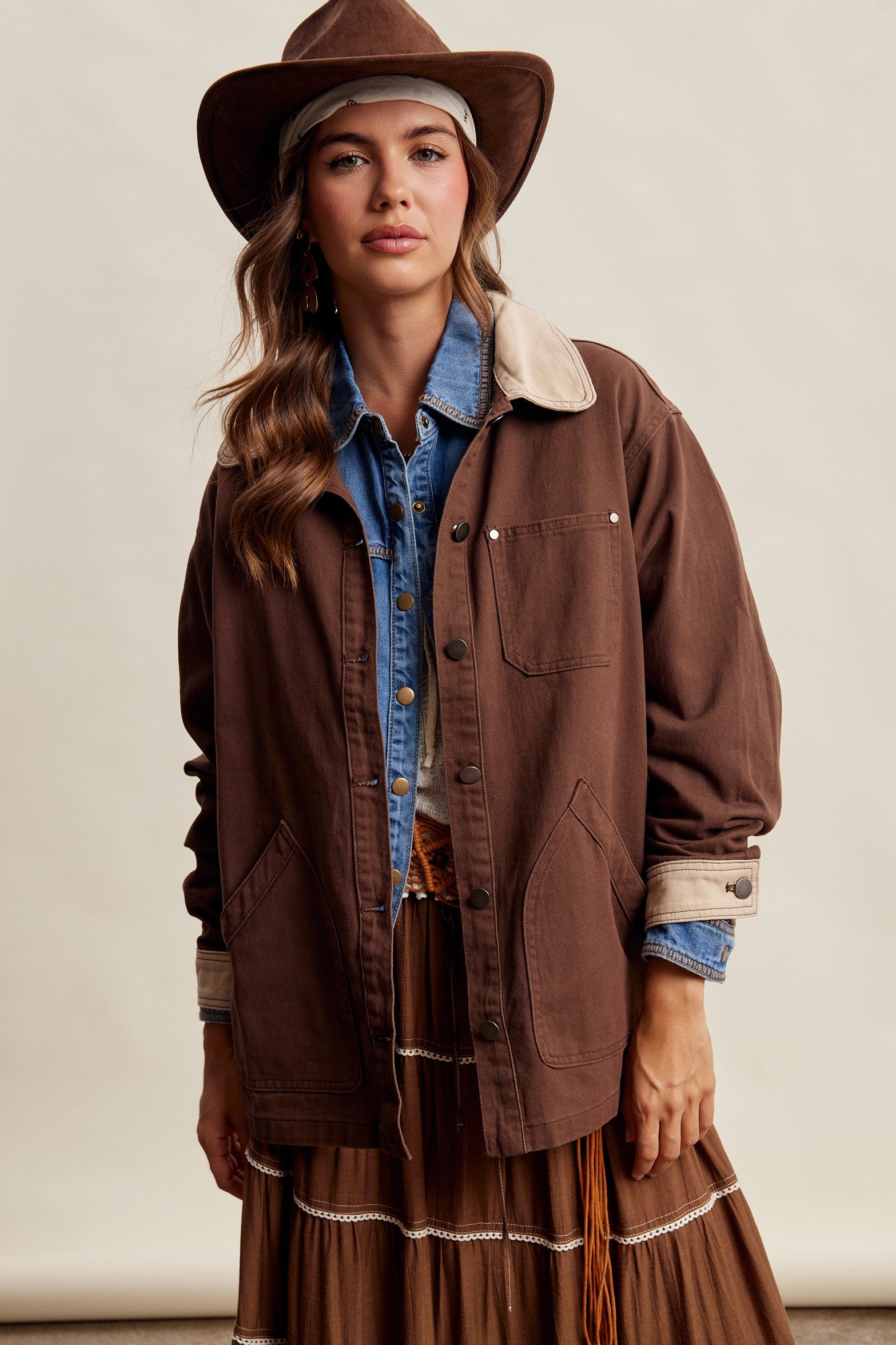 A woman wearing a brown wide-brim hat, a brown utility jacket with a light beige sherpa collar, a blue denim shirt, and a brown fringed maxi skirt, posed against a neutral beige studio background.