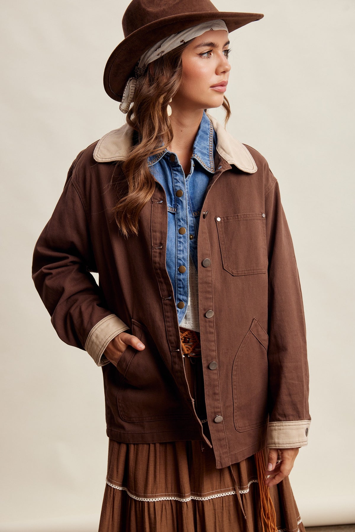 A woman wearing a brown  espresso colored button-front utility jacket with a light beige sherpa collar, styled with a blue denim shirt, brown hat, and brown skirt against a plain beige studio background.
