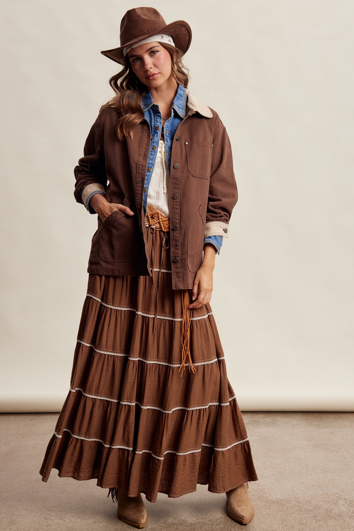 A woman modeling a brown espresso colored western-style outfit consisting of a brown utility jacket, matching pleated maxi skirt, blue denim shirt, white top, brown hat, and brown boots, against a plain beige studio backdrop.