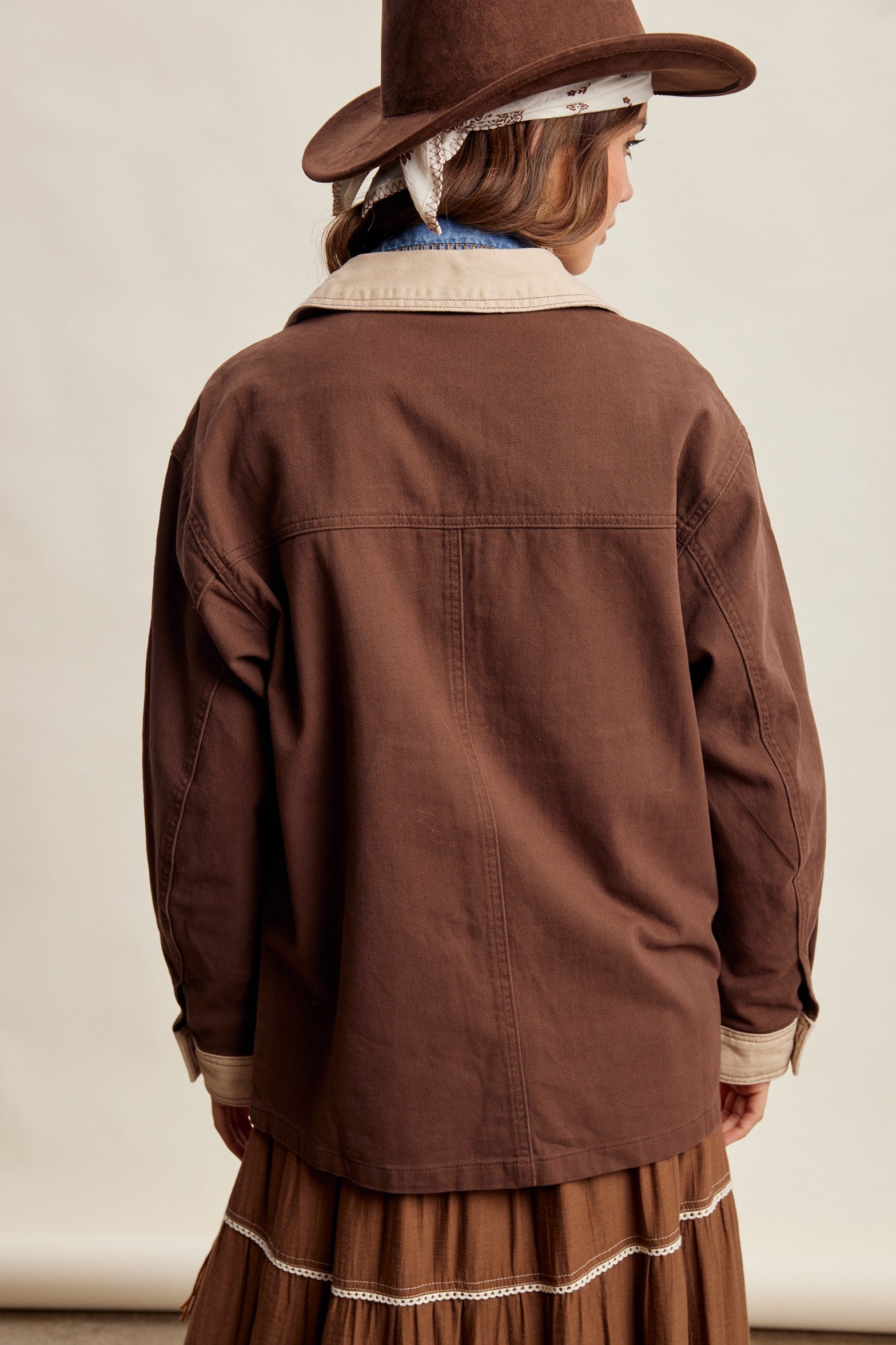 A woman wearing a brown wide-brim hat, a brown utility jacket with a light beige sherpa collar, a blue denim shirt, and a brown fringed maxi skirt, posed against a neutral beige studio background.