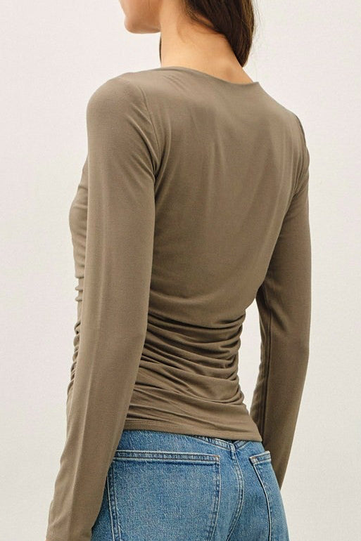 A woman shown from the back wearing a fitted olive long-sleeve top, blue jeans, and tan sneakers against a plain light gray studio background.