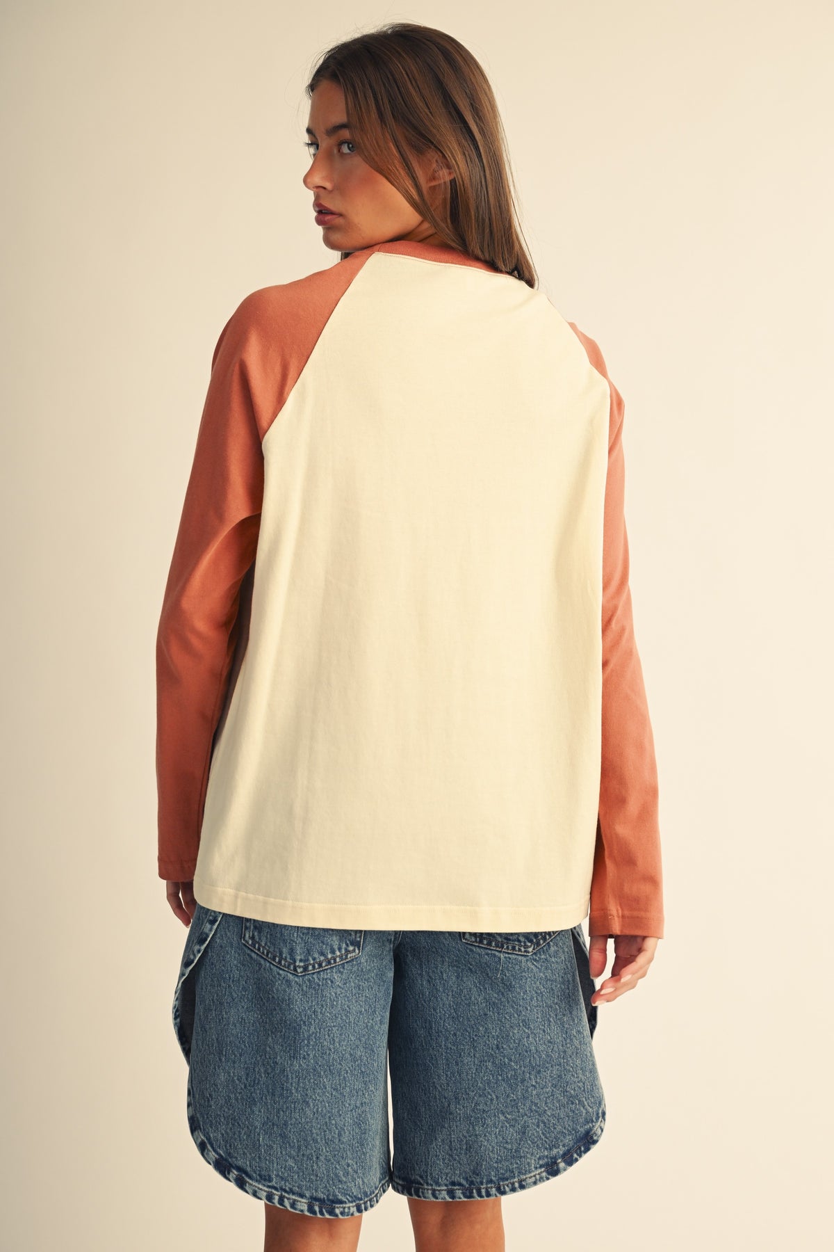 Back of women in a contrast raglan long sleeve baseball tee in butter terracotta and blue denim jean shorts.