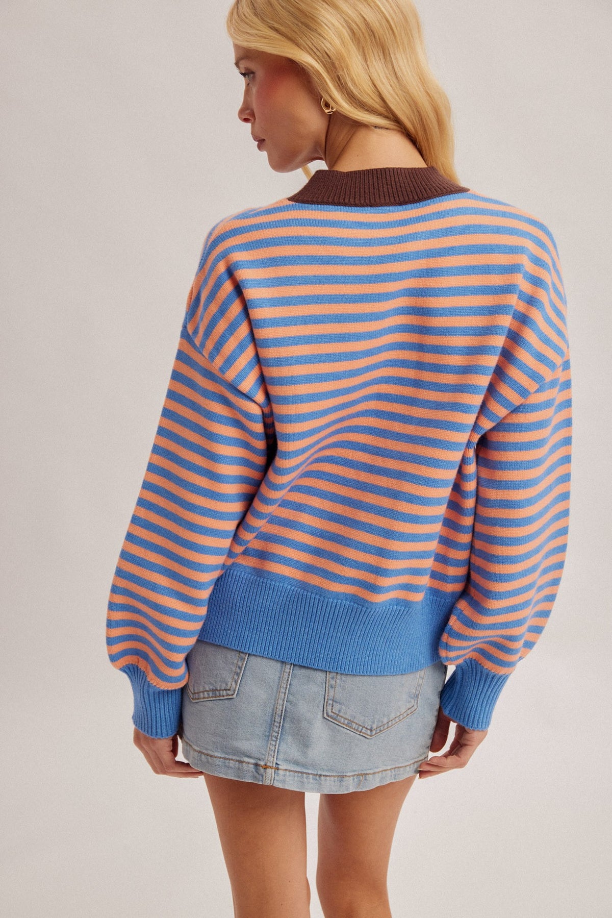A woman shown from the back wearing a blue and peach striped knit sweater with a brown ribbed collar, paired with a denim mini skirt, against a light gray studio background.