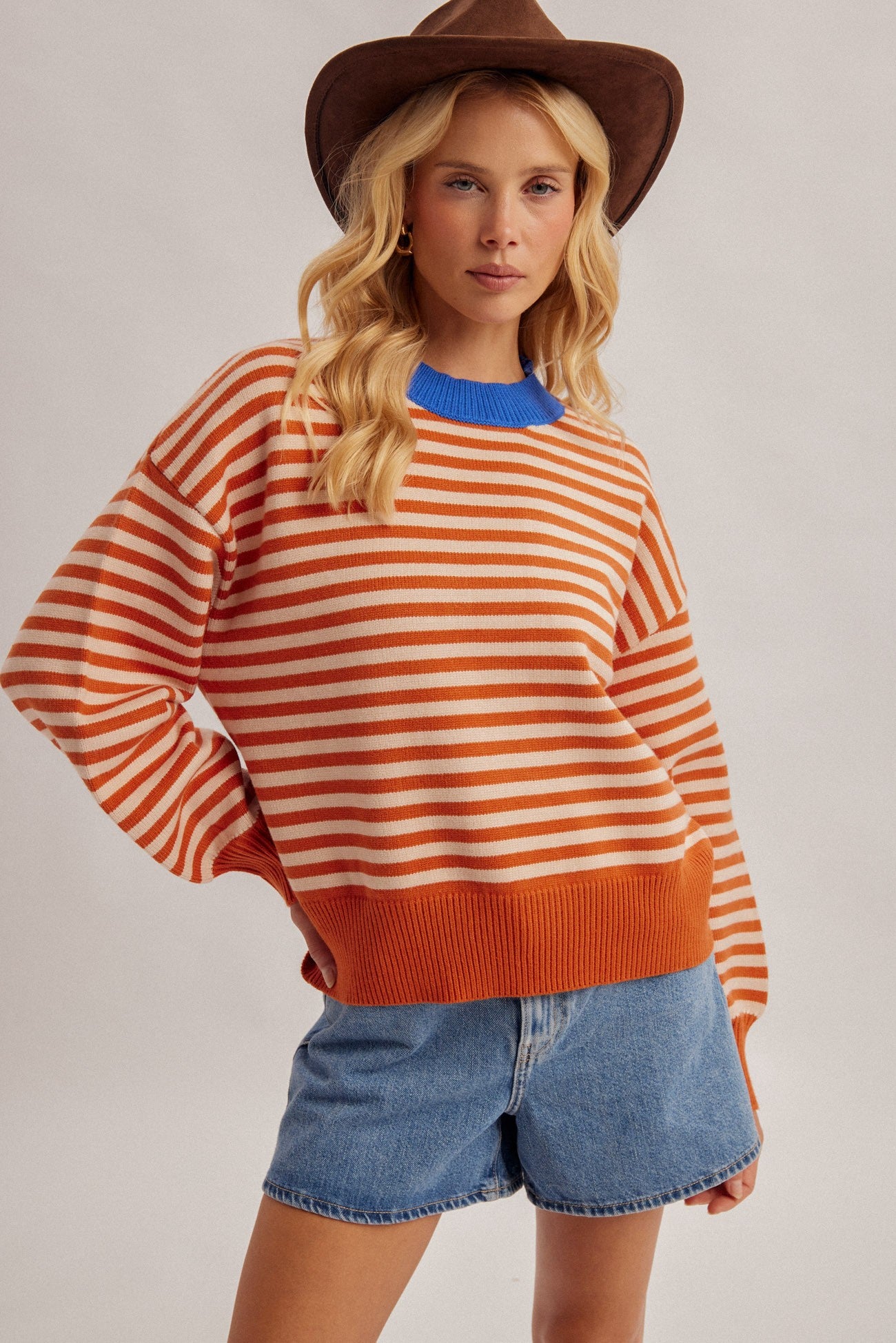 A women wearing a cowboy hat with a stripe pumpkin multi color sweater pullover with a denim skirt.