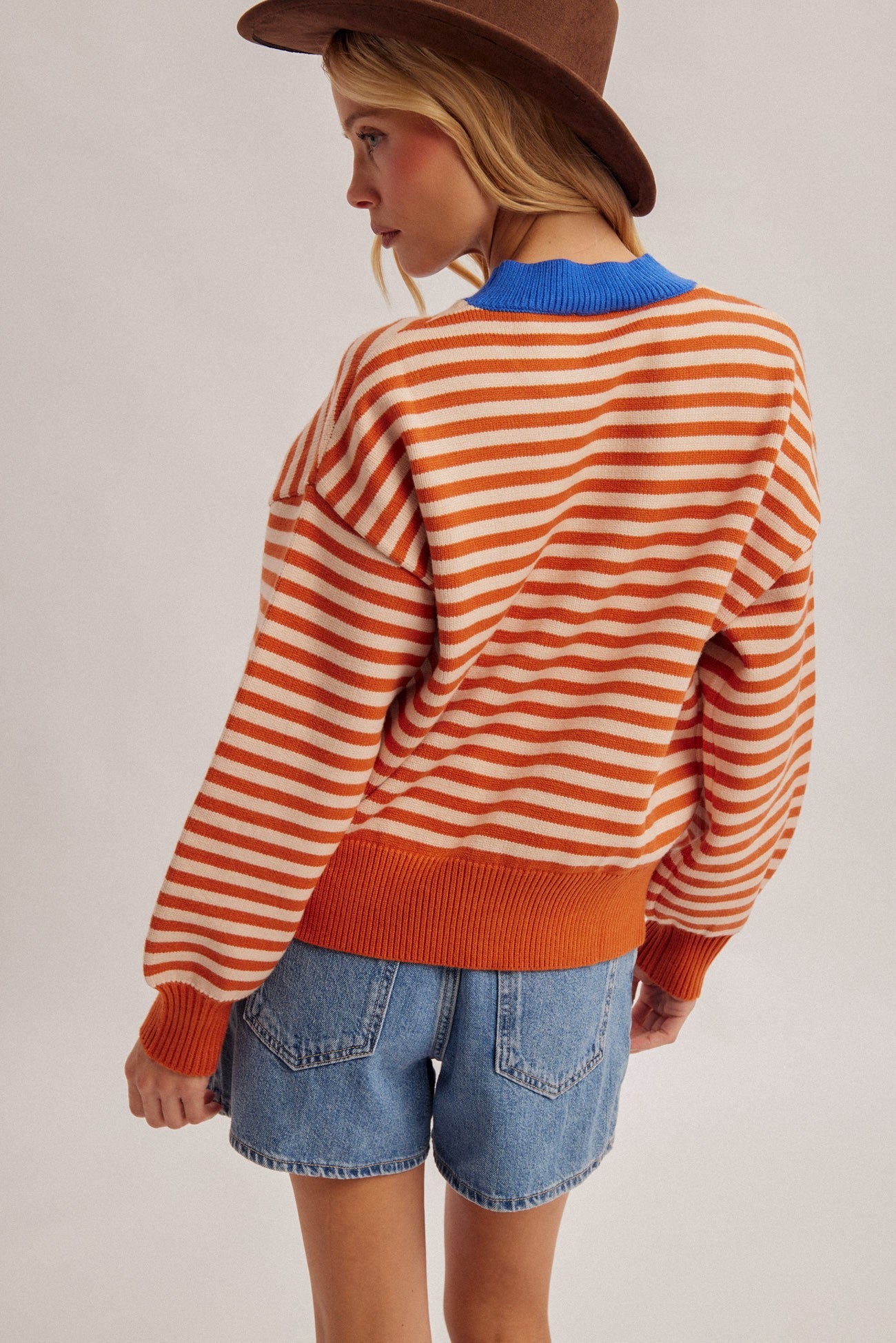 A women wearing a cowboy hat with a stripe pumpkin multi color sweater pullover with a denim skirt.