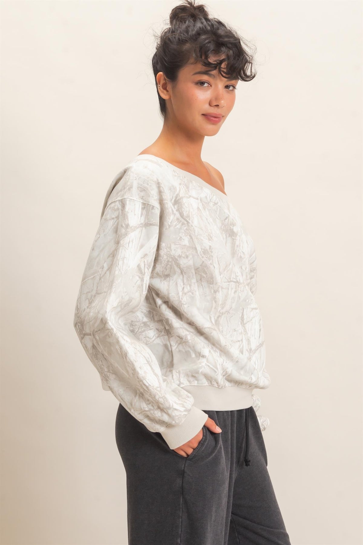 A woman modeling a textured, off-white long-sleeve sweater with a wide neckline, paired with dark gray sweatpants, against a plain beige studio background.