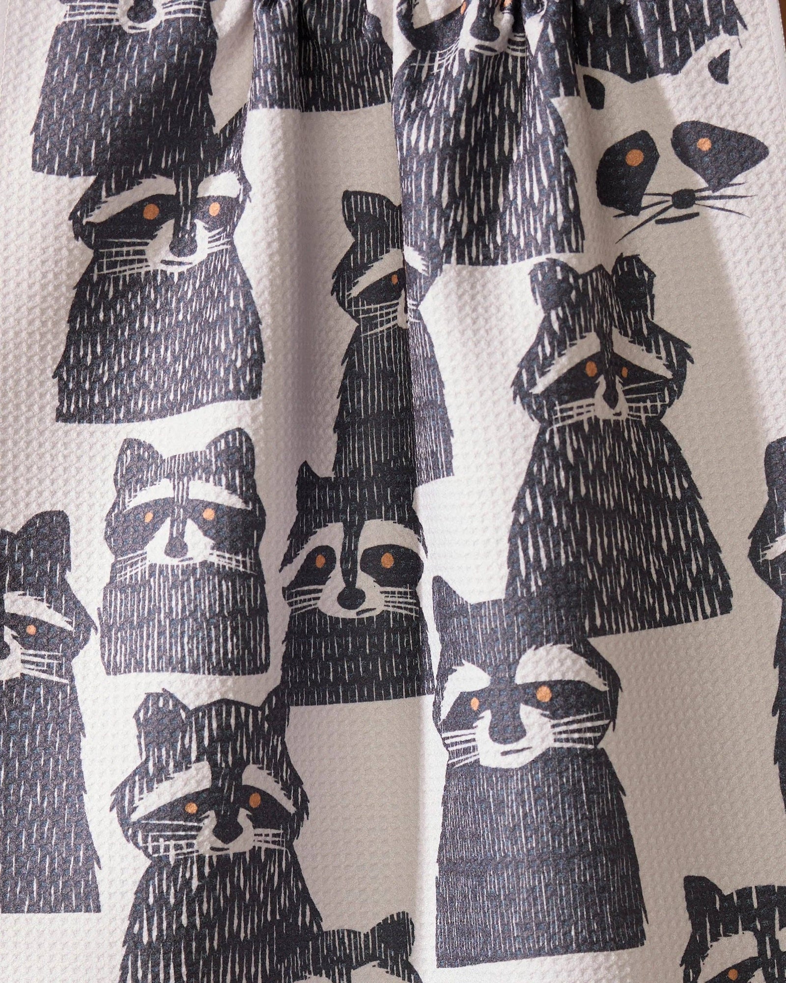 A white kitchen towel with a black raccoon pattern hangs from a string against a warm brown background, with a stack of white plates partially visible in the lower right corner.