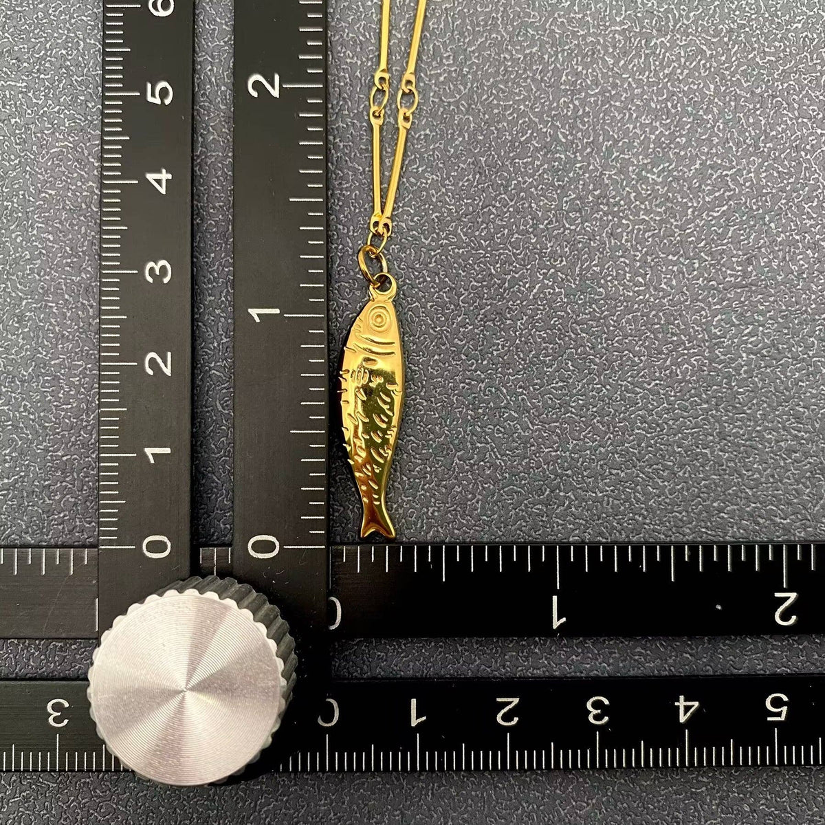 Gold fish-shaped pendant on a chain next to a ruler on a gray background