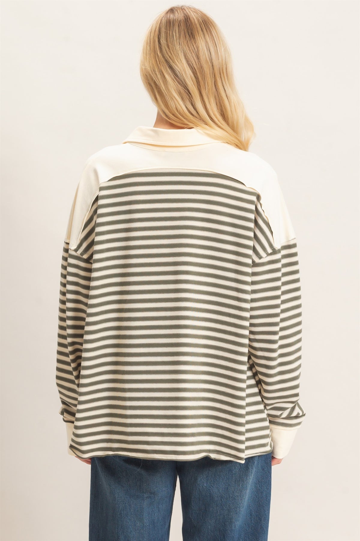 A woman viewed from the back wearing a long-sleeve olive gray top with horizontal stripes across the body and solid light cream sleeves and collar, paired with blue jeans against a plain light background.