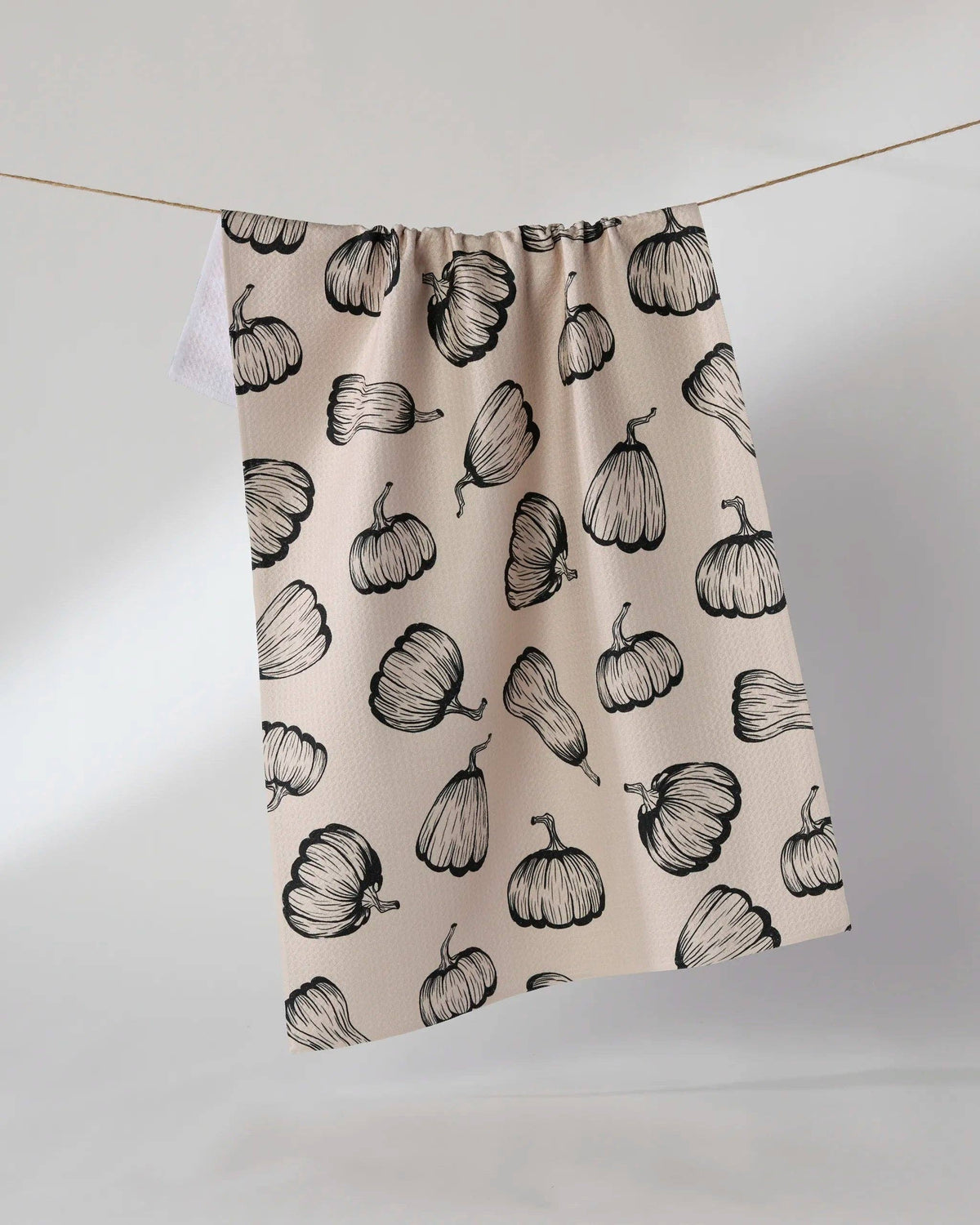 A beige tea towel with a black hand-drawn pumpkin pattern, hanging from a thin wire against a light gray studio background.