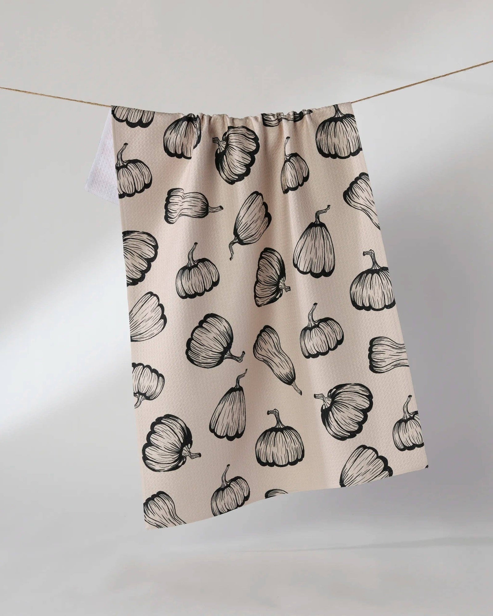 A beige tea towel with a black hand-drawn pumpkin pattern, hanging from a thin wire against a light gray studio background.