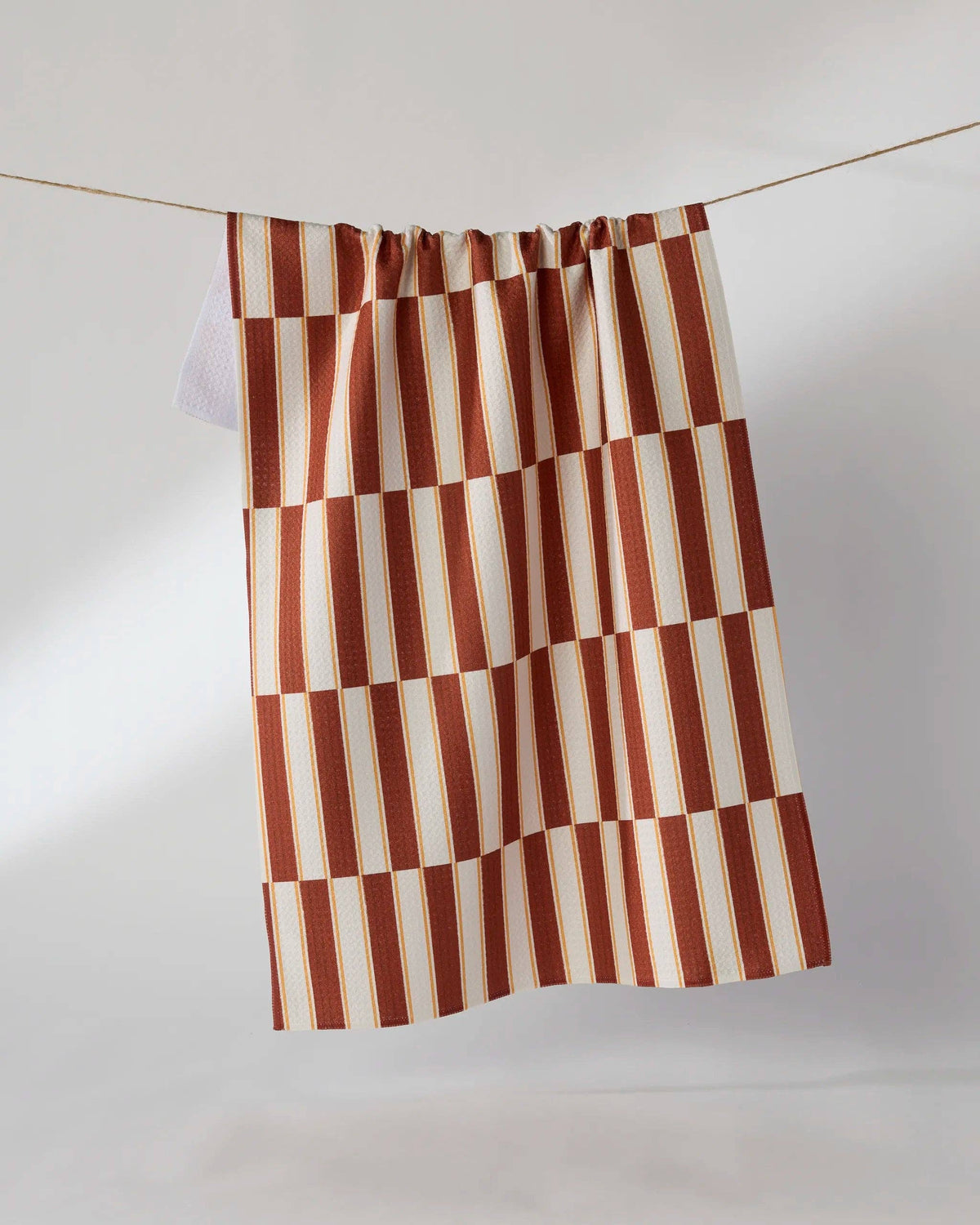 A tea towel with a rust red and cream checkerboard pattern hanging from a thin wire against a light gray studio background.