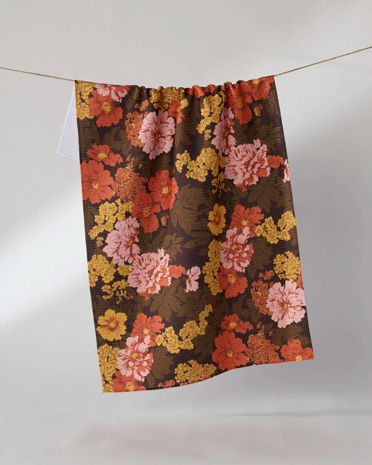 A rectangular fabric tea towel with a vintage floral pattern in orange, pink, and yellow on a dark brown background, hanging from a thin string against a light gray wall.