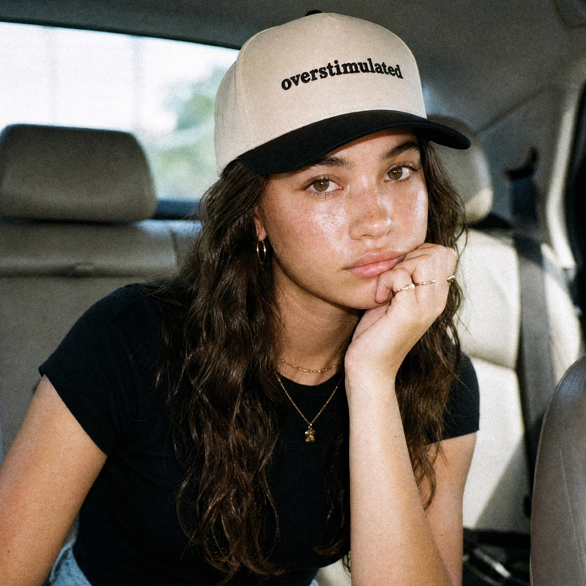 Girl wearing a hat that says overstimulated and a black tee.