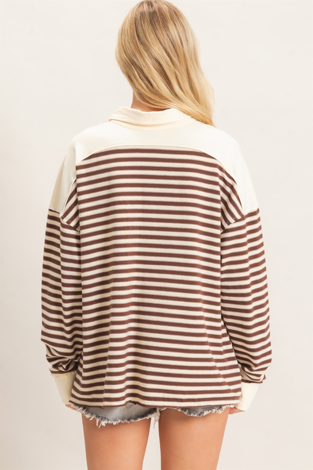 A woman viewed from the back wearing a long-sleeve knit top with horizontal brown and cream stripes and solid cream shoulder yokes, paired with denim shorts against a plain light background.