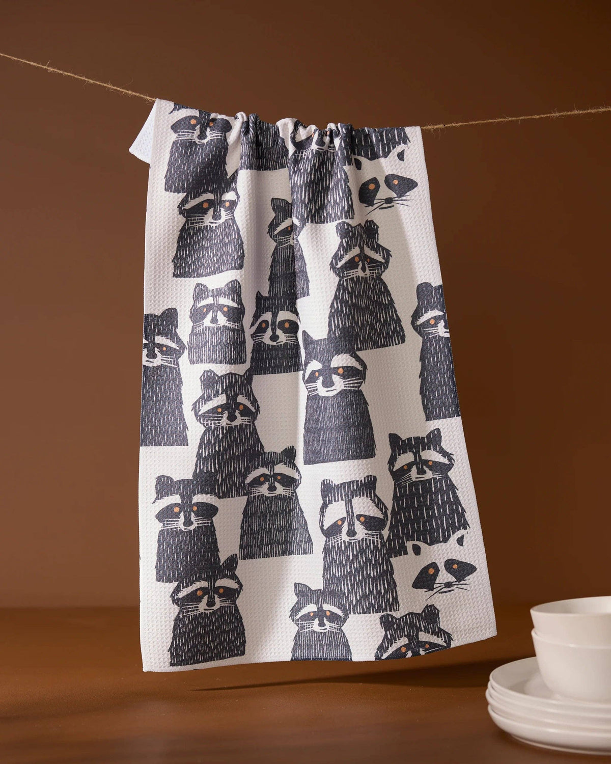 A white kitchen towel with a black raccoon pattern hangs from a string against a warm brown background, with a stack of white plates partially visible in the lower right corner.