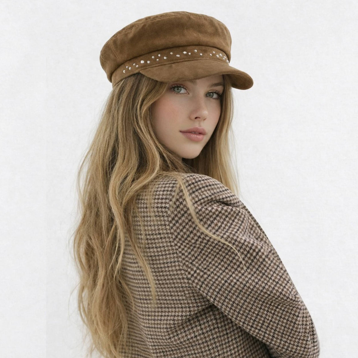 A woman with long blonde hair wearing a brown suede-like newsboy cap with a band of small white studs, paired with a brown and beige houndstooth jacket, photographed against a light gray background.