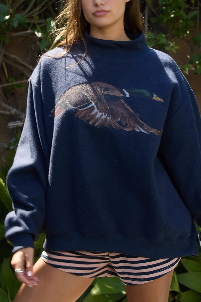 A woman wearing an oversized navy blue sweatshirt with a large mallard flying duck graphic on the front, paired with pink and navy striped shorts, standing outdoors in front of green foliage.