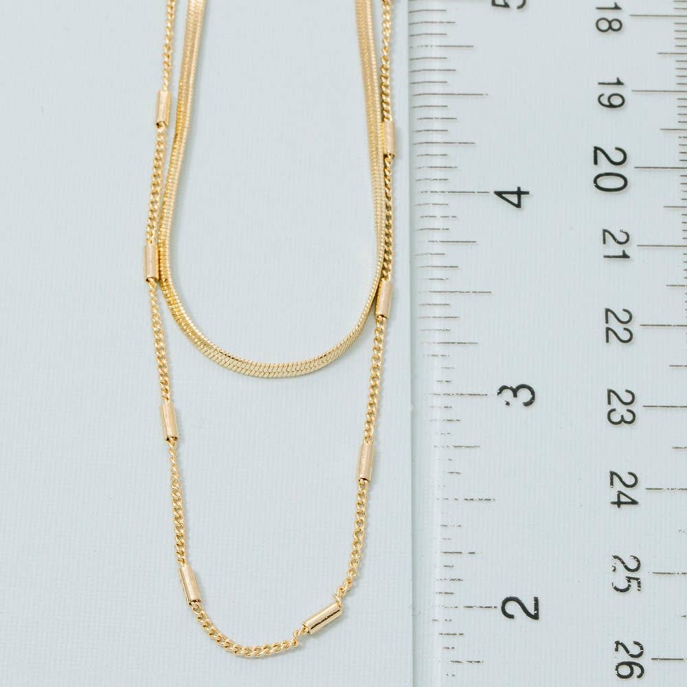 A gold-toned double-strand chain necklace with cylindrical links, displayed next to a white ruler for scale on a light blue background.