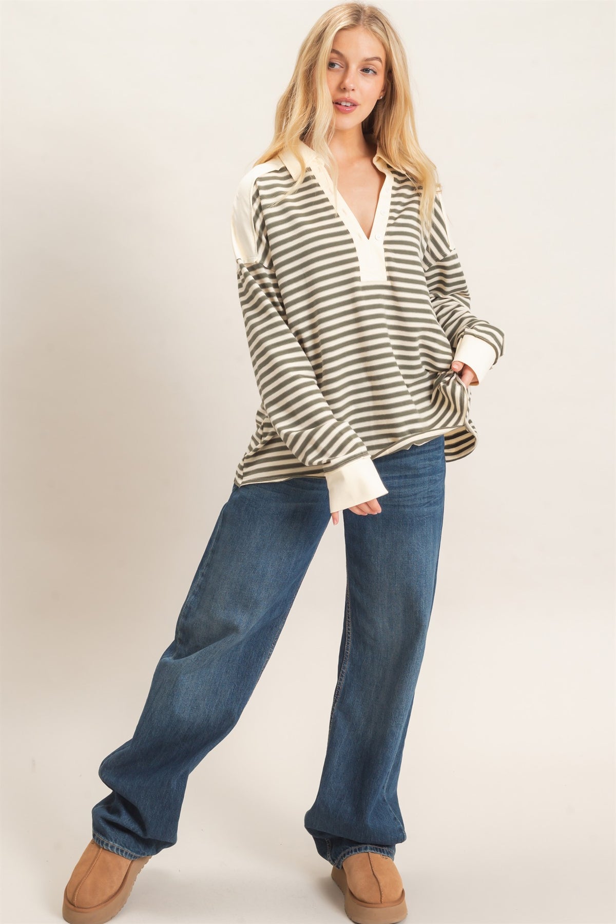 A woman modeling a striped olive gray top with wide sleeves and a V-neck, paired with loose blue jeans and tan shoes, against a plain light background.