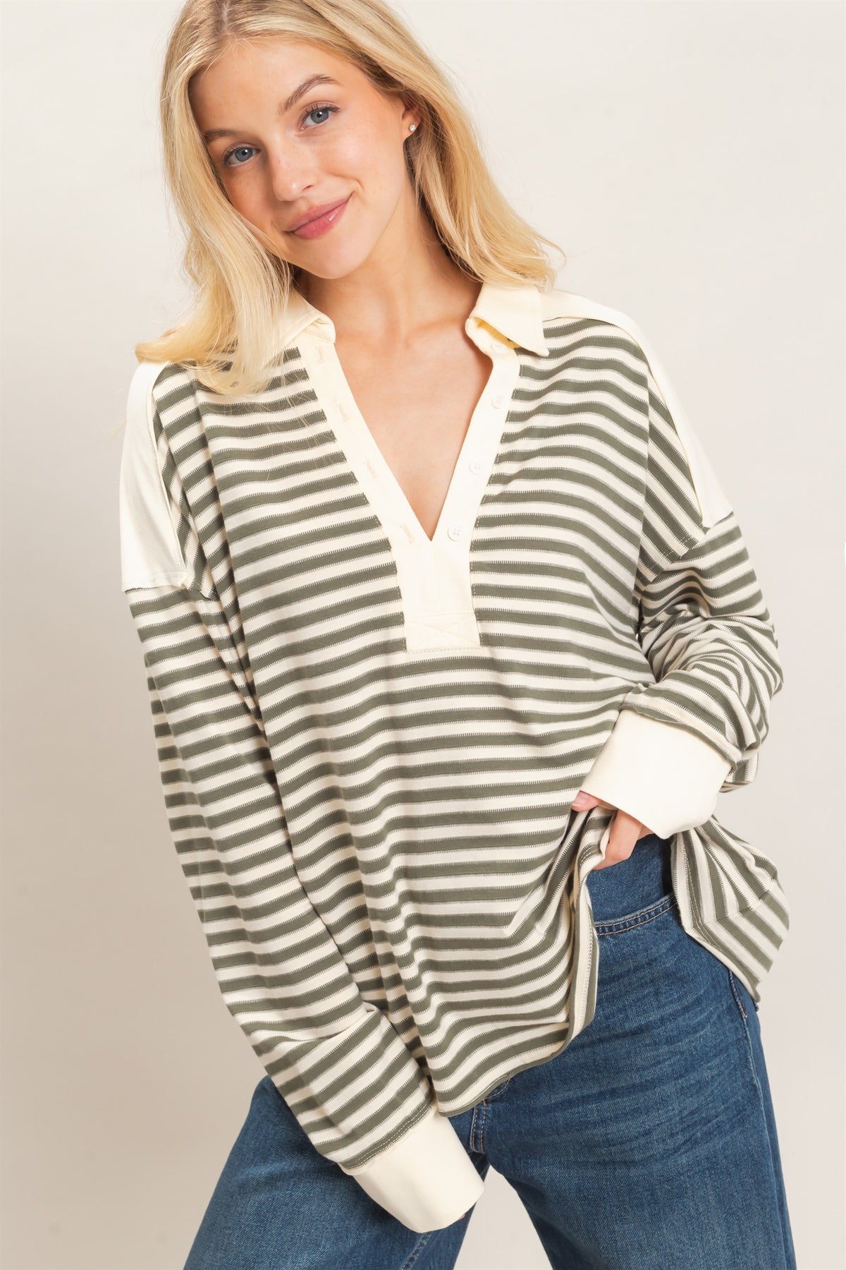 A woman wearing a long-sleeve collared knit top with horizontal olive gray green and cream stripes, paired with blue jeans, against a light beige studio background.