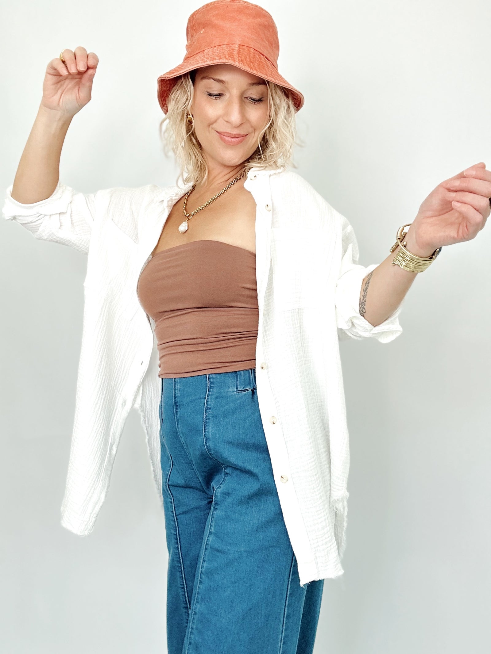 A woman modeling a gauze white button-up shirt over a brown bandeau top and blue jeans, wearing a terracotta bucket hat and gold jewelry against a light gray background.