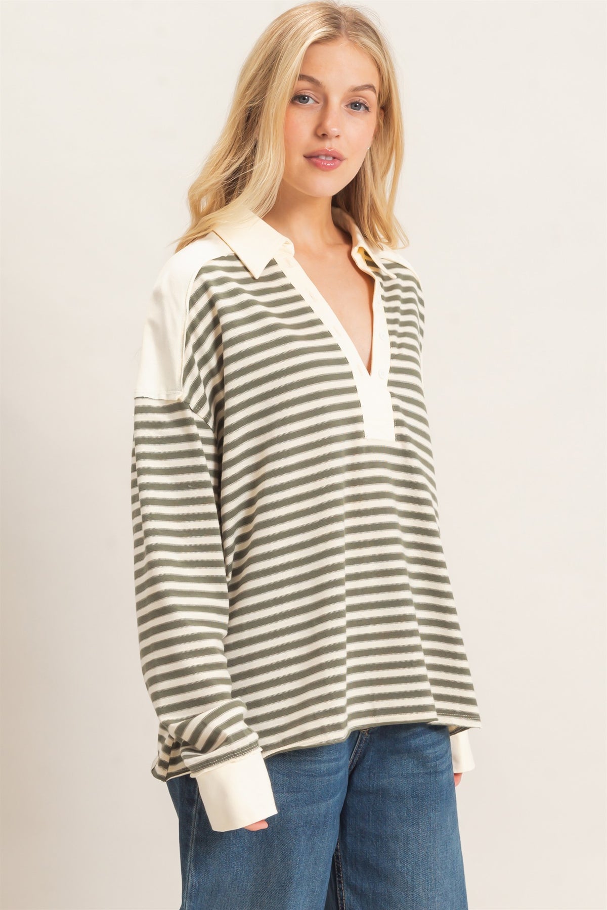 A woman wearing a long-sleeve collared knit top with horizontal stripes in olive gray green and cream, paired with blue jeans, against a plain light background.