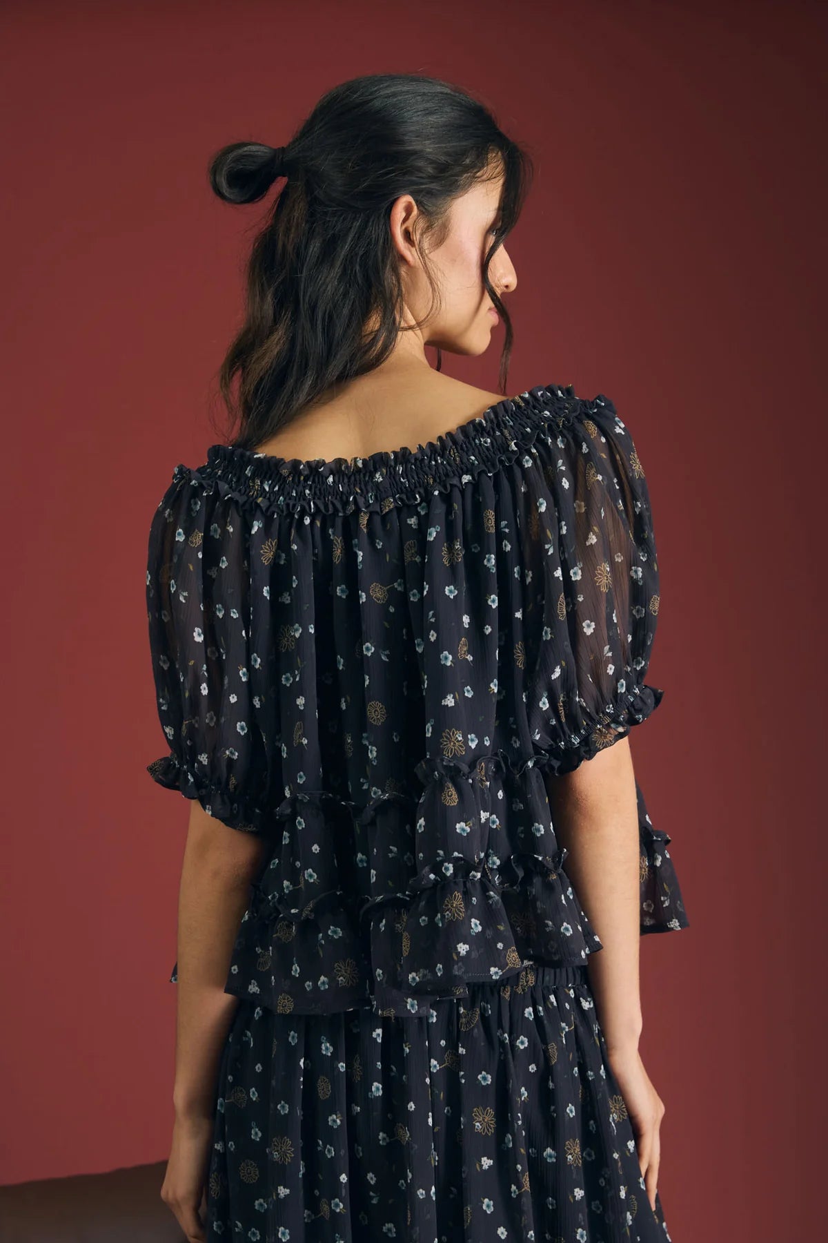 A woman shown from the back wearing a navy off-the-shoulder top with puff sleeves and a small floral print, set against a solid dark red studio background.