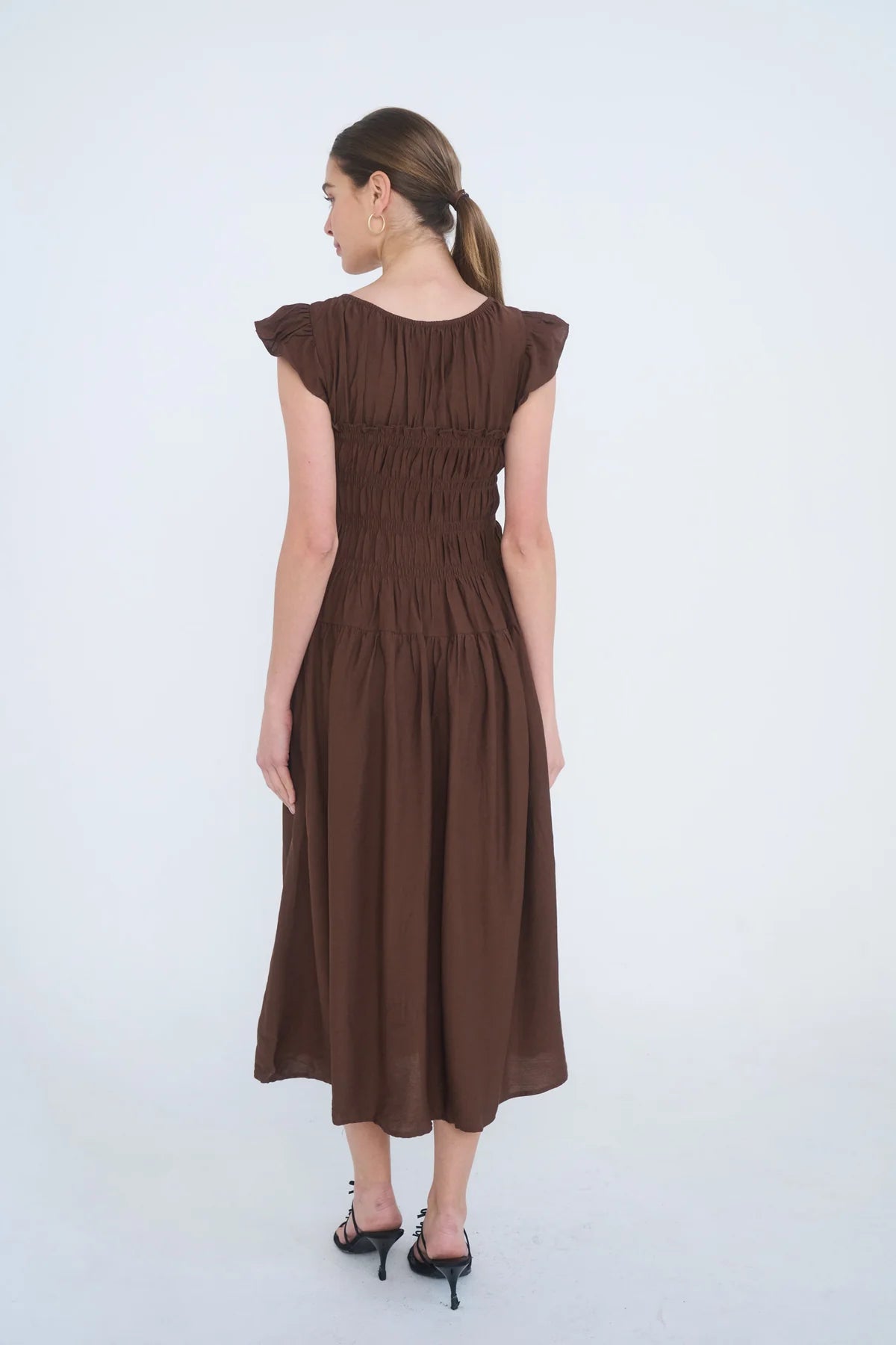 A woman wearing a brown midi dress with cap sleeves and a smocked bodice, shown from the back against a plain white studio background.
