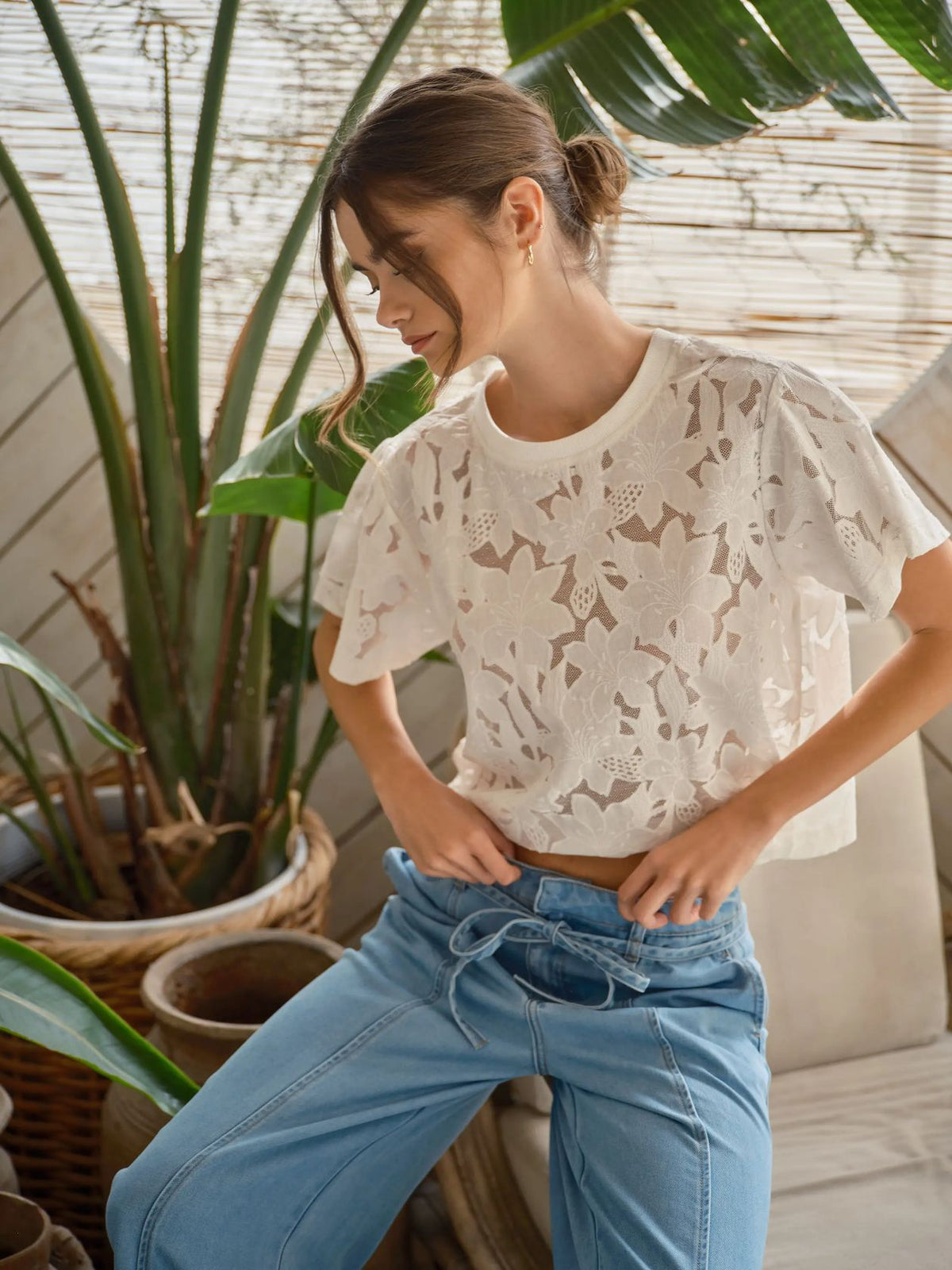 Woman wearing a white lace floral top and blue jeans standing in a room with plants.