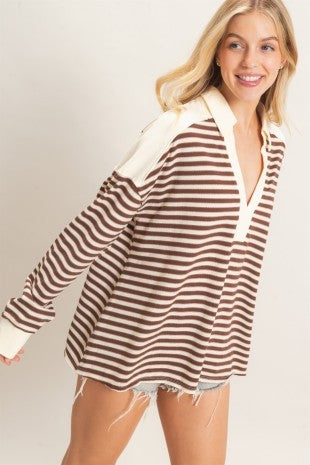A woman wearing a brown and cream striped long-sleeve top with solid cream shoulder panels and collar, paired with frayed denim shorts, against a light beige studio background.