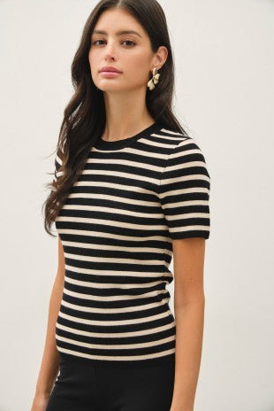 A woman wearing a short-sleeve crewneck top with horizontal black and cream stripes, paired with black pants, against a plain light gray background.